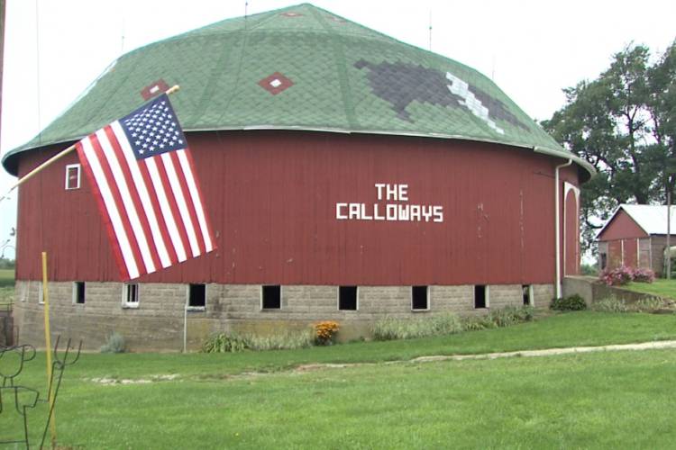 A red-colored round barn stands on grass, with the American flag flying next to it.