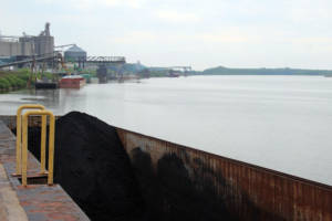 A barge of petcoke, a petroleum byproduct used to make aluminum, waits for offloading at the Port of Indiana-Mt. Vernon, with other barges carrying fertilizer, cement and soybean oil lined up on the Ohio River bank. (Annie Ropeik/IPB News)