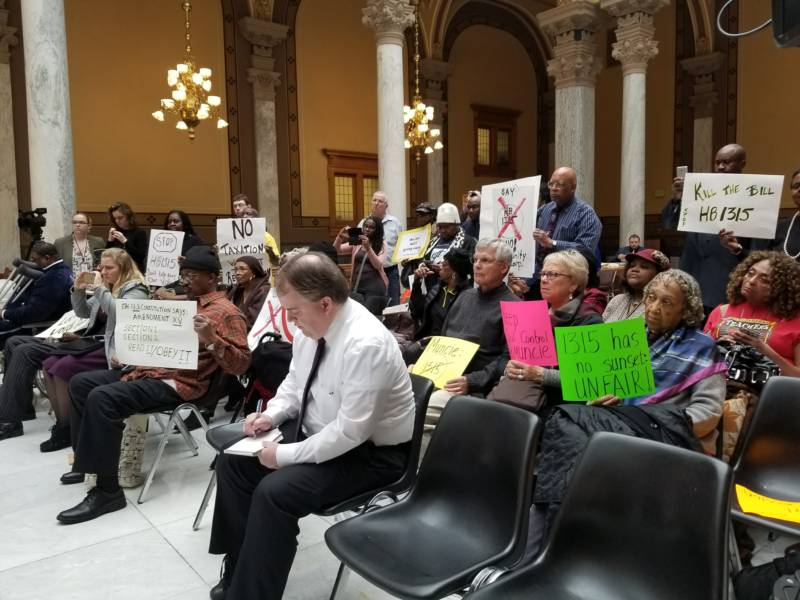 Protesters hold signs while seated in the audience of a Statehouse news conference.