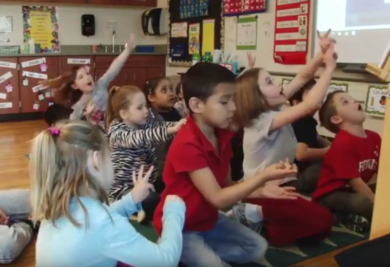 Elementary students sit on the floor, holding up their hands to answer the teacher's question.