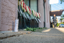 Flower memorials lay in front of a city building with a candle to the left and a police officer to the right background.