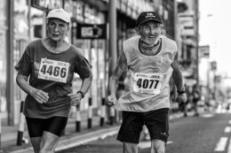 An elderly woman and man participate in a city marathon; they are wearing athletic clothes and numbered signs on their torsos. The man has a big smile on his face.