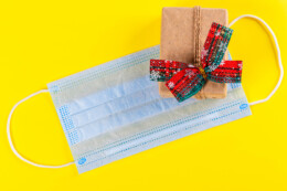 A face mask lays against a yellow background while a small, traditionally wrapped present sits on top of the mask.