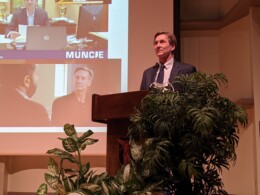 A white man stands behind a podium and potted plants, speaking into a microphone.