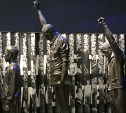 Gray statues in an exhibition depict the historical moment of The 'Black Power' Salute at the 1968 Olympic Games. The central statue is a man with his head lowered and his right arm raised with a fist.