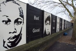 A wall is painted with black and white panels featuring portraits of people alternating with panels with the text "Bad" over "Good" or "Good" over "Bad." The panels alternate with black-on-white portraits and white-on-black text from the left foreground into the right background of the image. A tree's branches are visible over the right side of the wall.