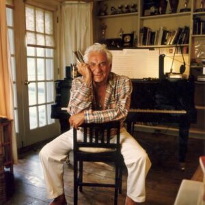 Composer and conductor Leonard Bernstein sits on a backwards chair in front of a piano. There are awards and books on his home shelf in the background.