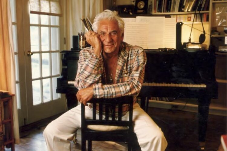 Composer and conductor Leonard Bernstein sits on a backwards chair in front of a piano. There are awards and books on his home shelf in the background.