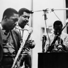 A black and white photo of John Coltrane playing the tenor sax, Cannonball Adderley playing the alto sax, and Miles Davis playing the trumpet in a studio.