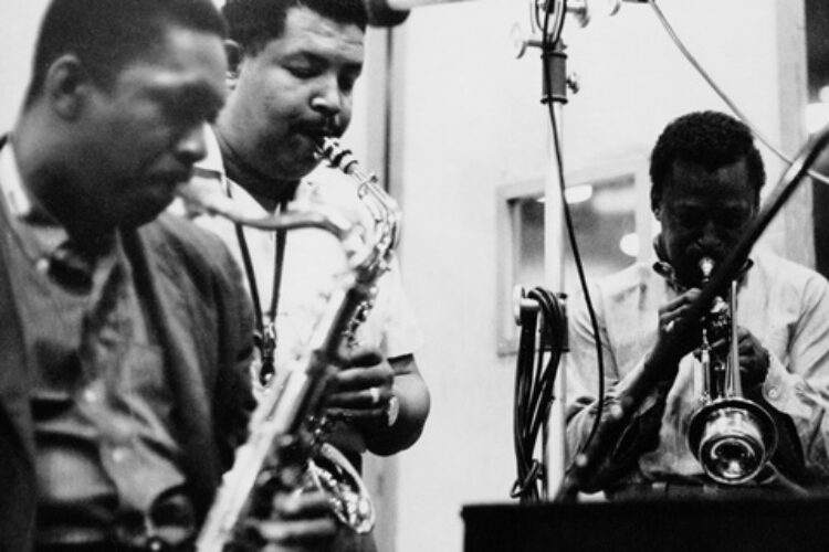 A black and white photo of John Coltrane playing the tenor sax, Cannonball Adderley playing the alto sax, and Miles Davis playing the trumpet in a studio.