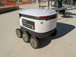 A delivery robot that's white in color, with six wheels.