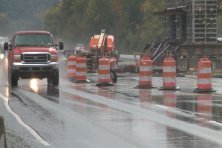 A red truck drives down a road in the rain. To the left of the truck is orange construction cones and road work equipment