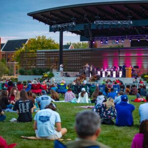 A band plays in a small outside amphitheater. Dozens of listeners sit in the grassy area directly in front if it