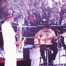 Jazz saxophonist Sonny Rollins plays saxophone next to a sitting electric bassist onstage during a music performance. We can see the audience in the background.