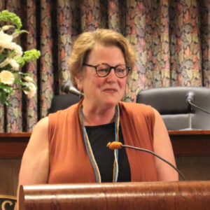 A woman with short blond hair and glasses stands at a podium, speaking and smiling.