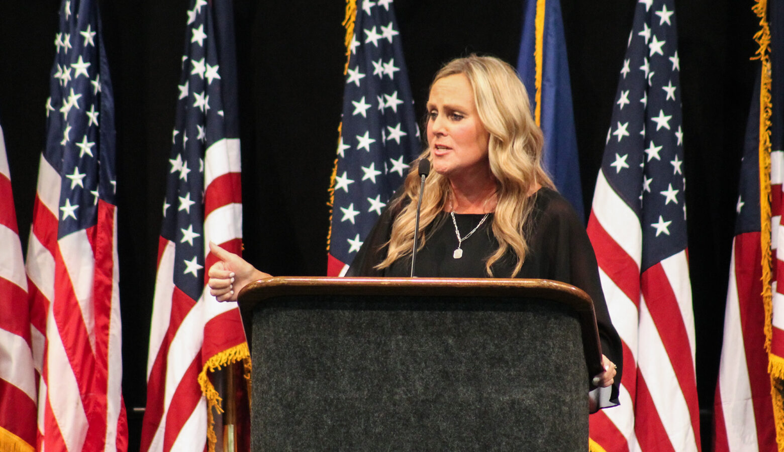 Jennifer McCormick speaks at a podium at the Indiana Democratic State Convention. Behind her is a black curtain with a line of American flags. McCormick is a White woman with blonde hair. She is wearing a black top, with a necklace and large pendant.