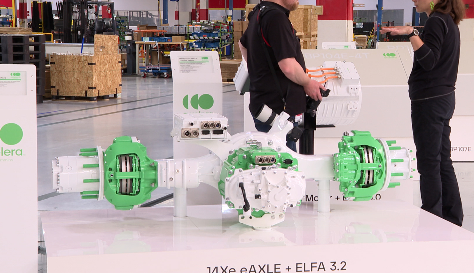 An electric vehicle powertrain prototype is displayed at an event. The machine is white and green and sits on a platform with the label 14Xe eAXLE ELFA 3.2 on its side. Two people wearing mostly black are speaking to one another in the background of the image.