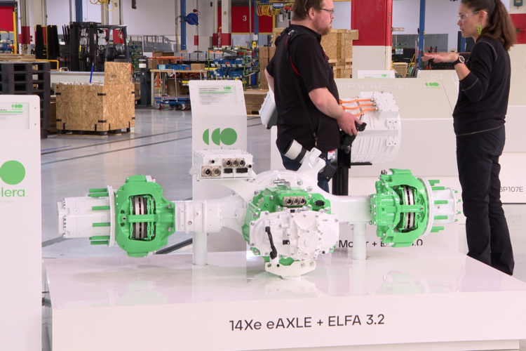 An electric vehicle powertrain prototype is displayed at an event. The machine is white and green and sits on a platform with the label 14Xe eAXLE ELFA 3.2 on its side. Two people wearing mostly black are speaking to one another in the background of the image.