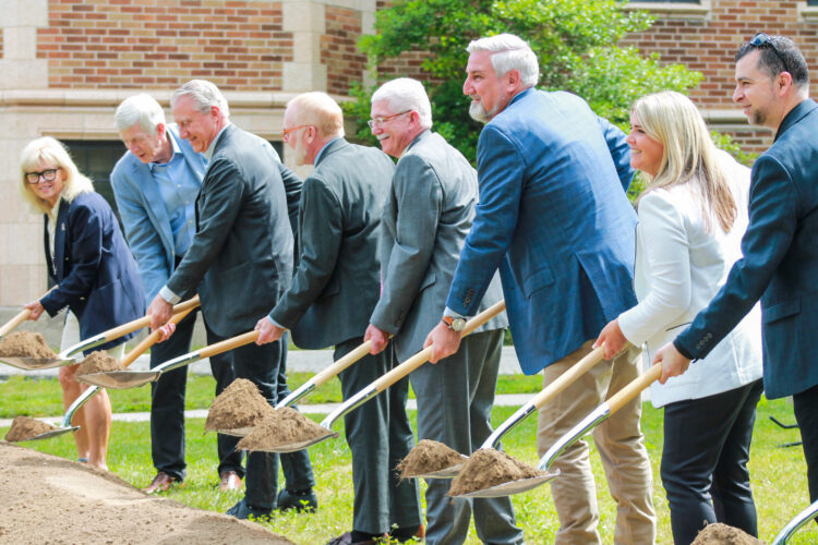 A row of people in suits hold shovels loaded with dirt over a mound.