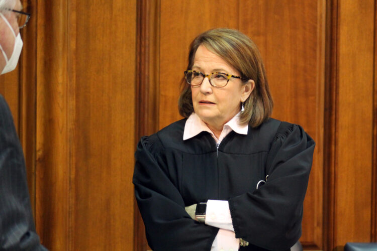 Loretta Rush stands with her arms folded while speaking to someone on the Indiana House floor. Rush is a White woman with brunette hair. She is wearing glasses and black judicial robes over a light-colored shirt.