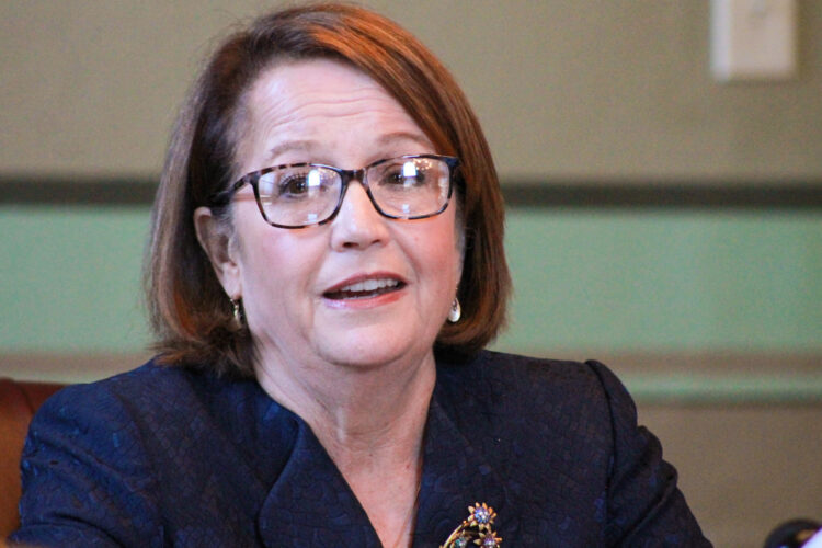 Loretta Rush speaks while sitting at a table. Rush is a White woman with brunette hair. She is wearing glasses and a blue jacket.