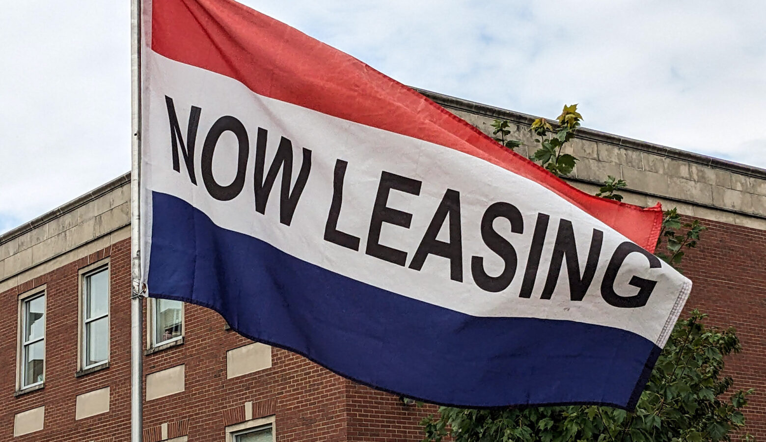 A flowing flag with a red stipe on the top, a blue stipe on the bottom, and a white stripe in the middle that has the words "now leasing" in block lettering.