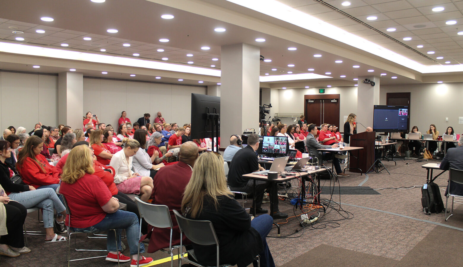 People in mostly red shirts sit in a chairs at a State Board of Education meeting.