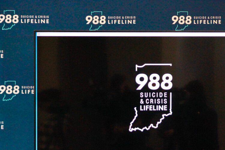 A large black screen has the 988 hotline logo in its center. Behind it is a blue backdrop with the 988 hotline logo stamped across it repeatedly.