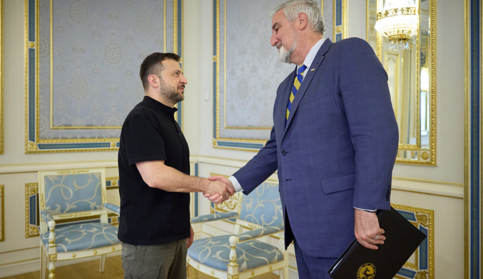 Eric Holcomb is a White man with white and gray hair and beard. He shakes hands with Ukrainian President Volodymyr Zelenskyy, who has dark hair and a short beard.