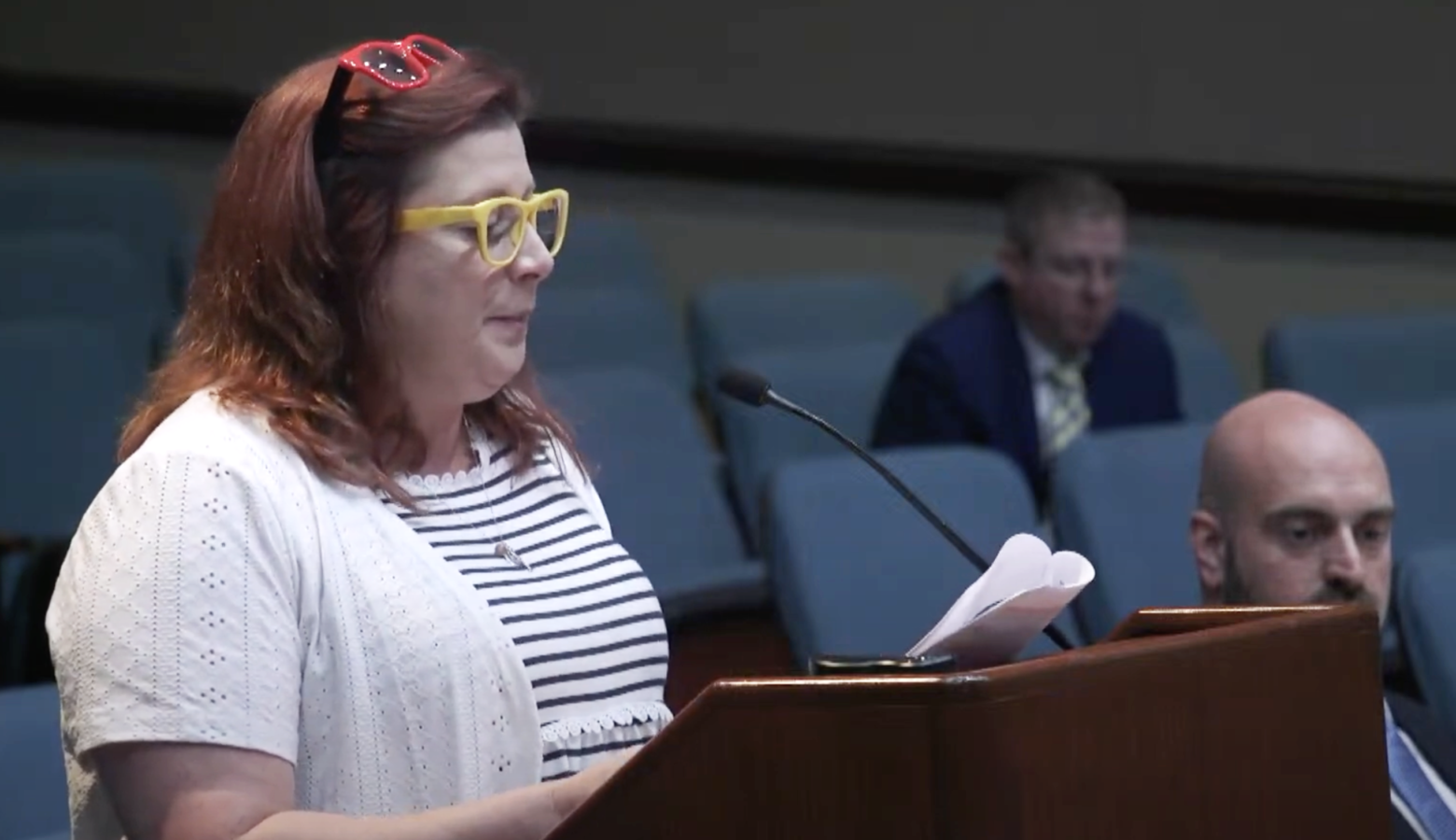 Abby Means stands at a podium holding a packet of papers that have been stapled together. Means is a White woman with shoulder length red hair.