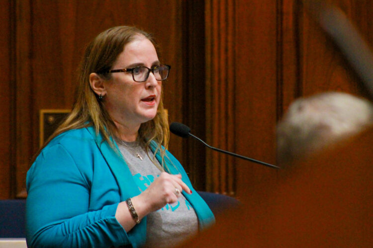 Amber Davis testifies in the Indiana House. She is speaking into a microphone at a lectern. Davis is a White woman with light brown hair. She is wearing glasses and a teal sweater over a gray t-shirt.