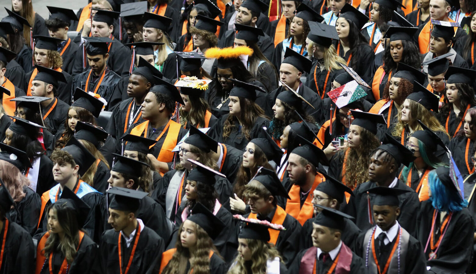 Dozens of high school graduates are wearing their black caps and gowns inside of an arena.