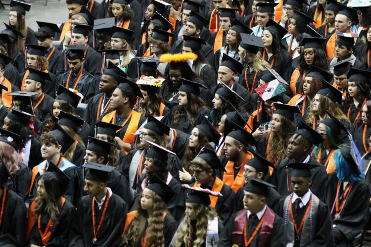 Dozens of high school graduates are wearing their black caps and gowns inside of an arena.