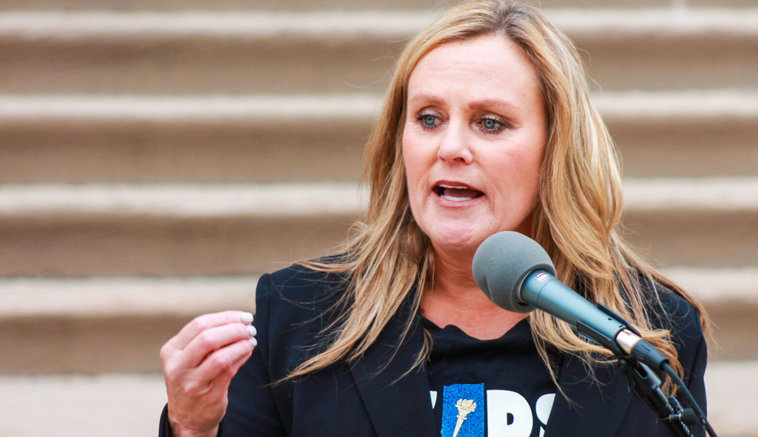 Jennifer McCormick speaks into a microphone outside the Statehouse. McCormick is a White woman with blonde hair. She is wearing a black jacket over a black t-shirt with a logo of the state of Indiana.