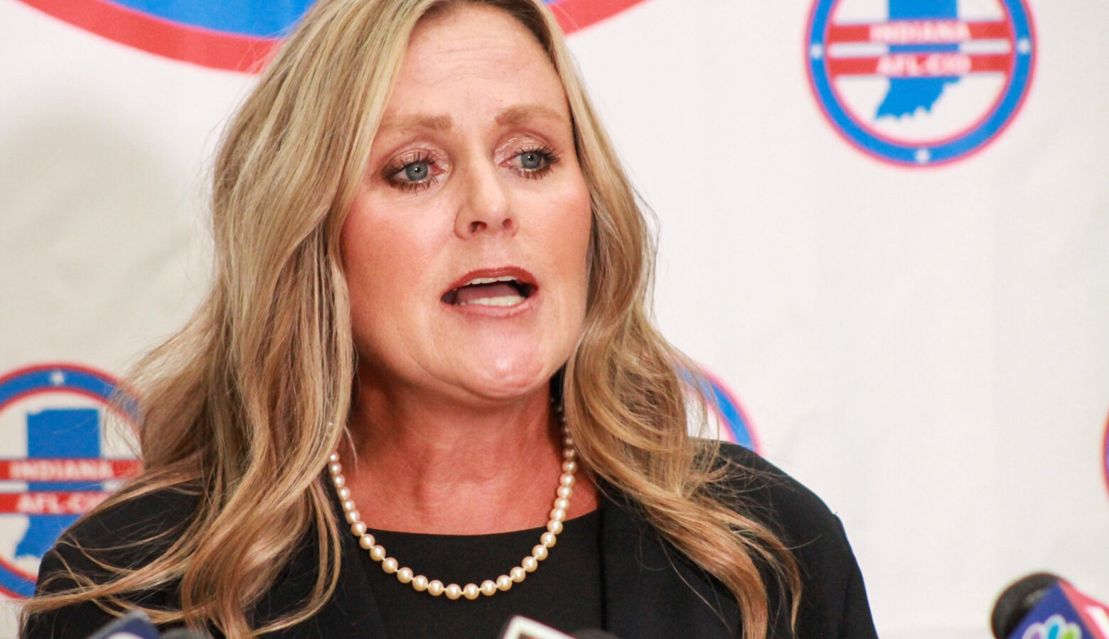 Jennifer McCormick speaks into a bank of microphones during a press conference. McCormick is a White woman with blonde hair. She is wearing a black jacket over a black top.