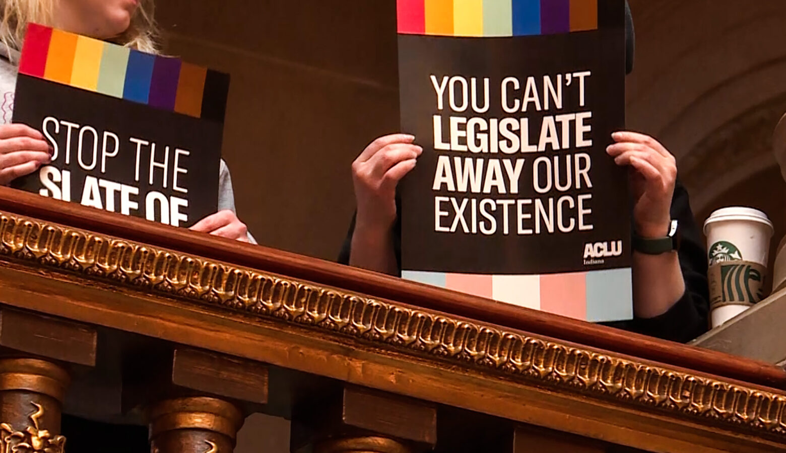 Protesters at the Statehouse hold up signs that read "You Can't Legislative Away Our Existence" with the ACLU of Indiana logo.