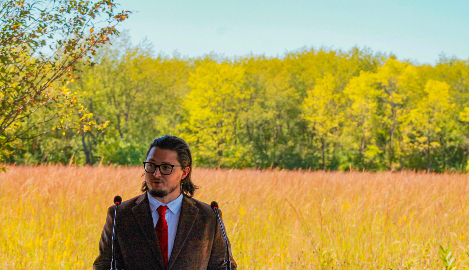 Logan York stands at a lectern in front of prairie grass and, further in the background, a forest. York is a man with long, dark hair and a beard. He is wearing glasses and a brown sport coat over a shirt and tie.