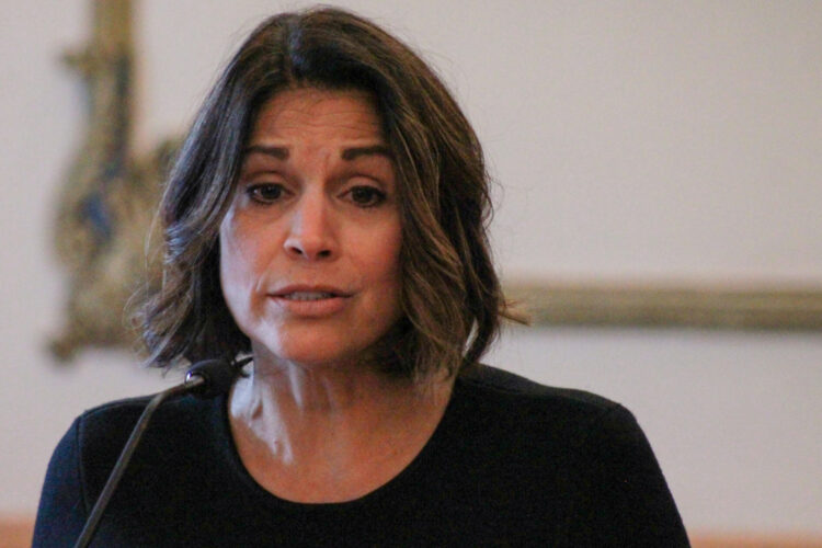 Mary DeBoer speaks into a microphone. DeBoer is a White woman with dark hair. She is wearing a black top.