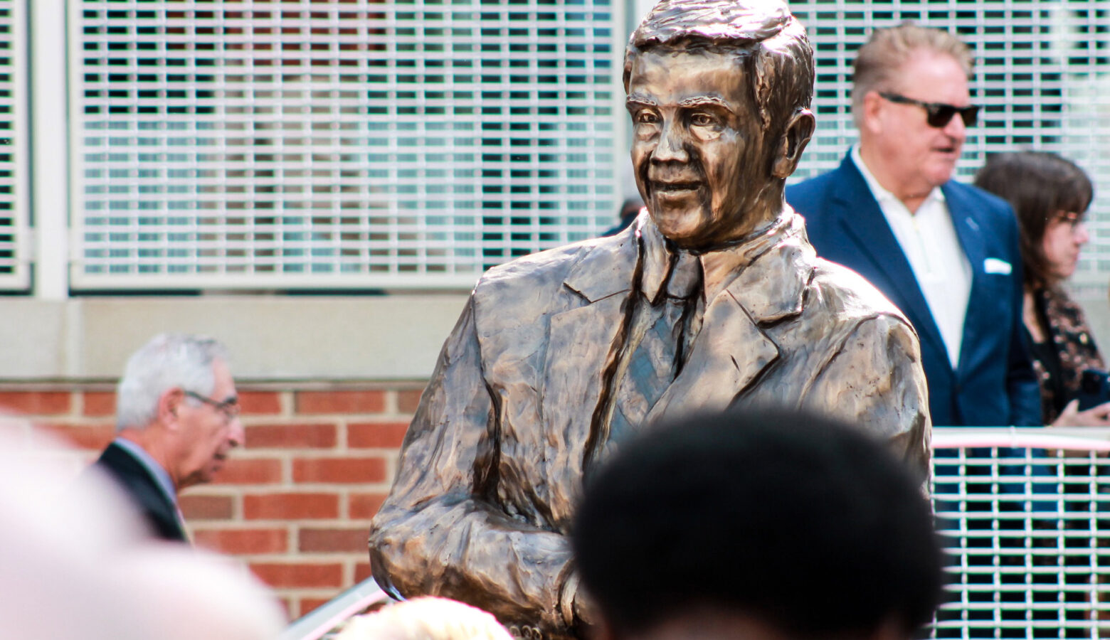 A statue of Richard Lugar stands above a crowd of people. The statue is in bronze. It shows Lugar wearing a suit and tie and leaning on a pedestal.