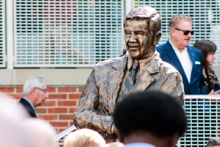 A statue of Richard Lugar stands above a crowd of people. The statue is in bronze. It shows Lugar wearing a suit and tie and leaning on a pedestal.