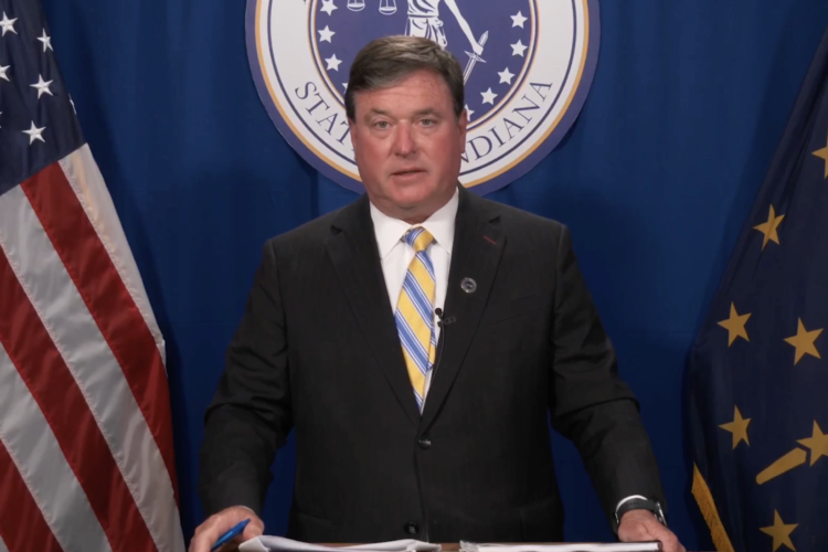 A screenshot of a Facebook Live shows Todd Rokita standing at a lectern in front of the American and Indiana flags, with the seal of the office of the attorney general behind him. Rokita is a White man with dark, graying hair. He is wearing a suit and tie.