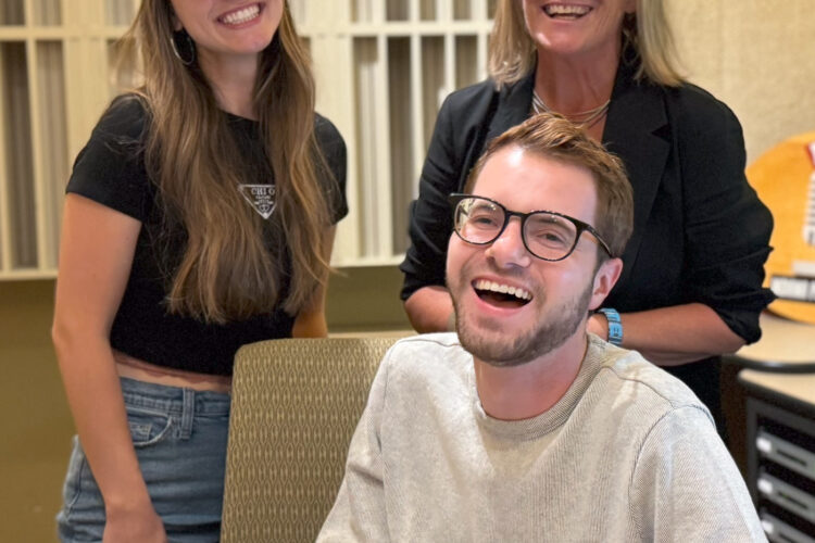 Three people stand together, with one sitting at a desk.