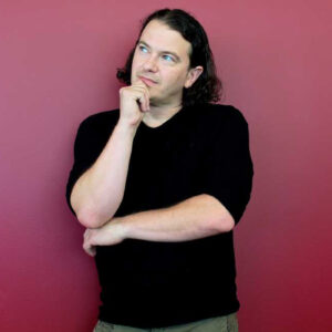 A man in a black t-shirt leans against a red wall with a thoughtful expression and pose.