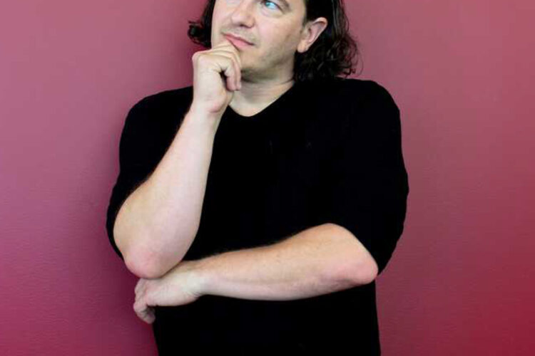 A man in a black t-shirt leans against a red wall with a thoughtful expression and pose.