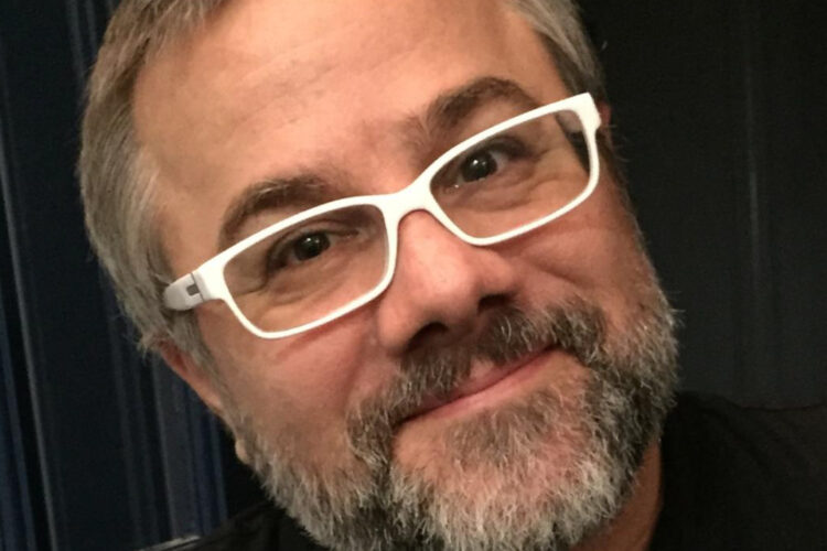 A man with a salt-and-pepper beard and rectangular white glasses smiles at the camera.