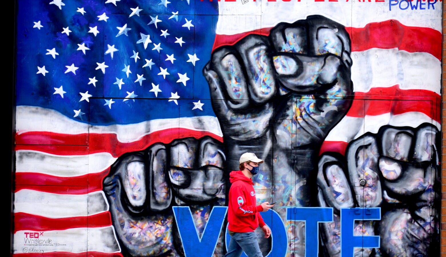 A person in a red hoodie walk past a wall mural. It depicts three raised fists in front of the American Flag with the word "vote."