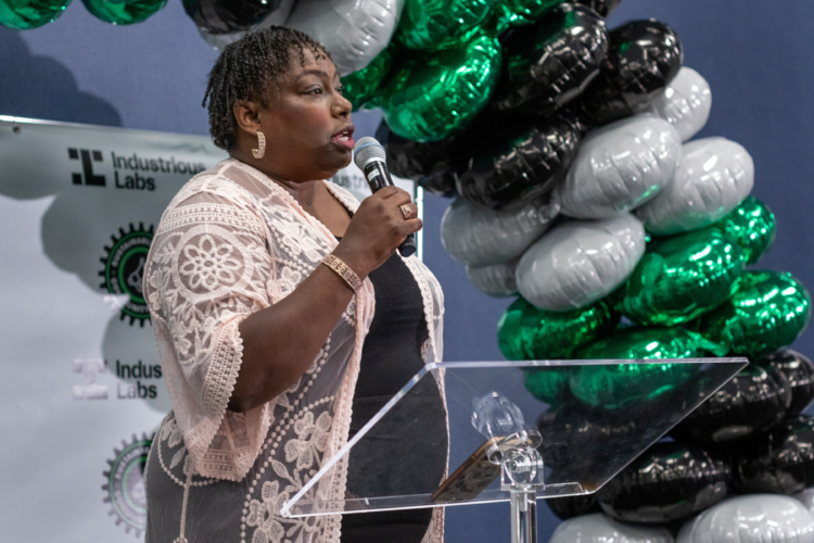 Beverly Lewis holds a microphone at an event. She is a Black woman with short hair, and stands in front of a balloon arch.