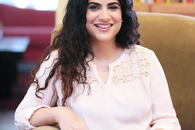 A smiling woman with long, wavy black hair sits on an armchair.