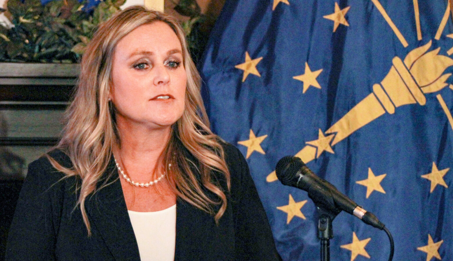 Jennifer McCormick speaks into a microphone with the Indiana flag draped behind her. McCormick is a White woman with blonde hair. She is wearing a black jacket over a white top.