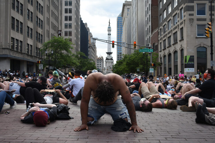 Several dozen protesters lay on the brick roads in downtown Indianapolis as part of a "die in." In the foreground, the main subject of the image is a Black protester, on their hands and knees, shirtless, with their head hung. Directly behind them is Indianapolis's Monument Circle.